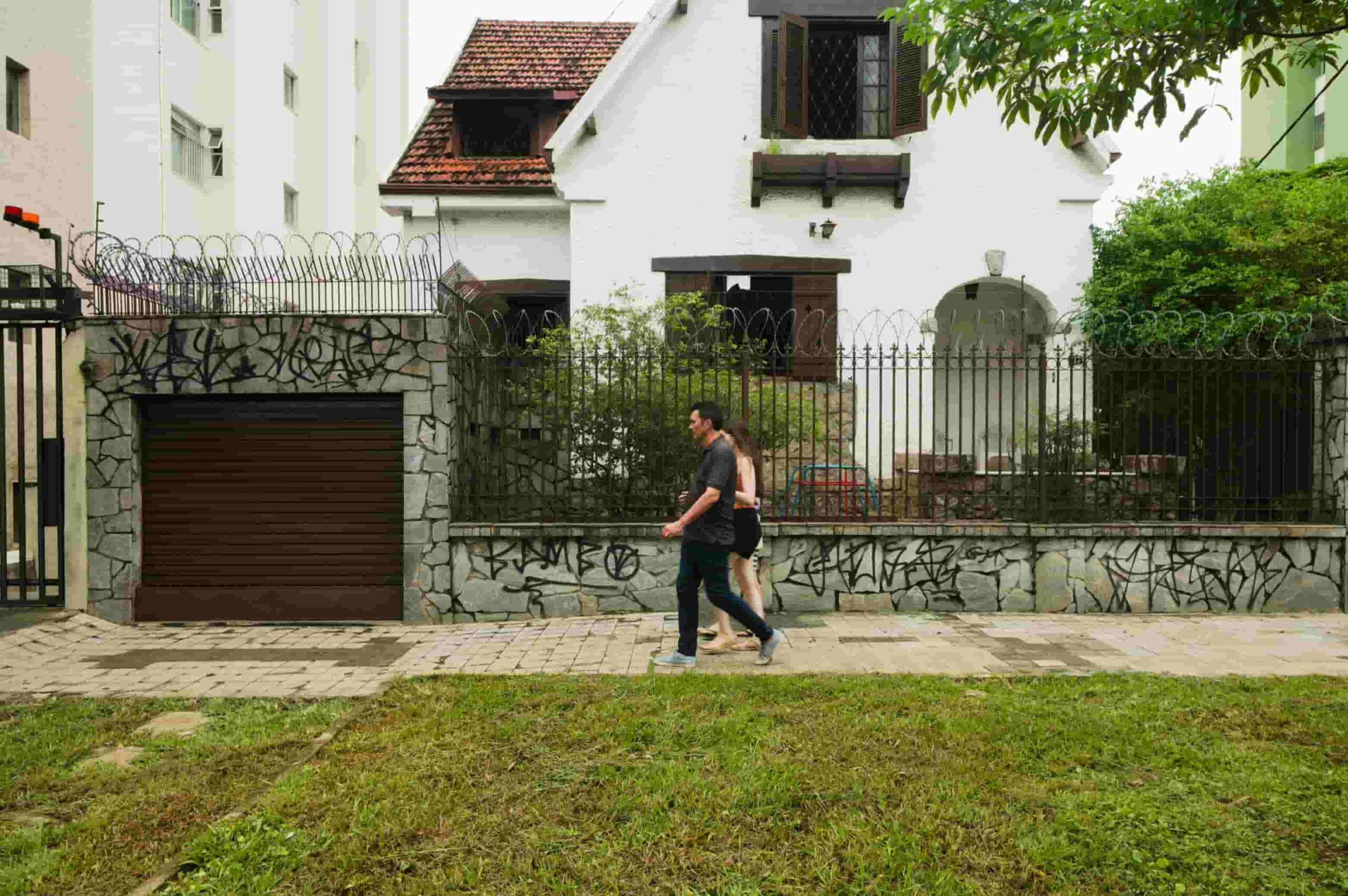 Two people walking on a sidewalk in front of a residential house with a tiled roof and surrounding greenery.