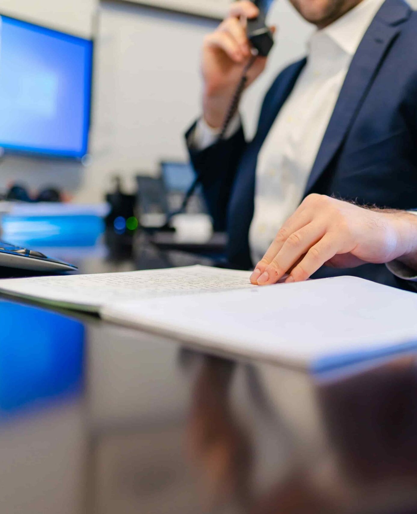 Person in a suit speaking on the phone while reviewing documents at an office desk.