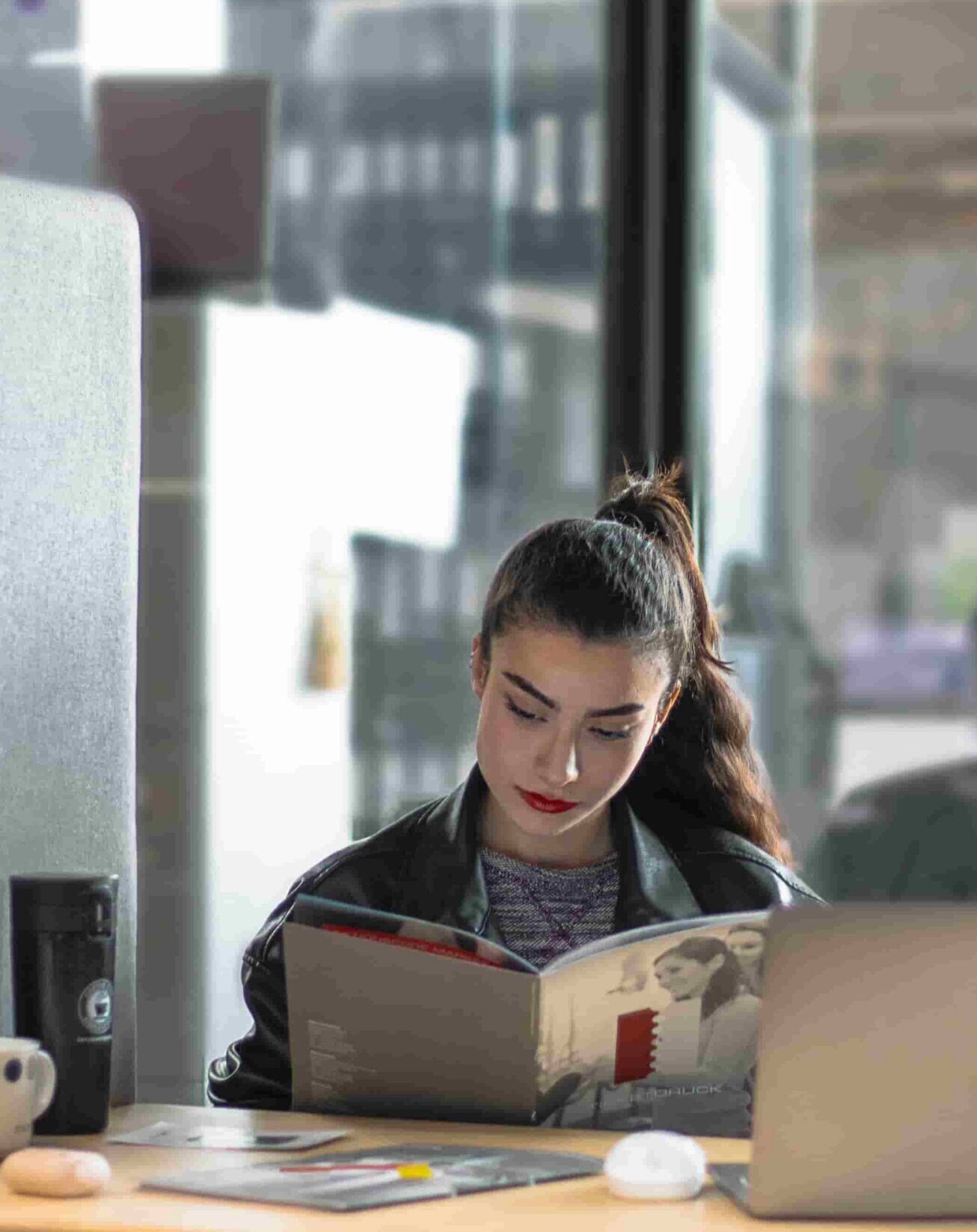 Woman sitting at a office desk reading a booklet beside a laptop in a bright coworking space.